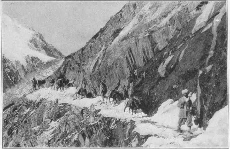 A row of men and horses walking along a snowy path on the side of a mountain.