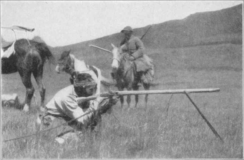A man aiming down the barrel of a long rifle. In the background are horses, one of which is carrying another man.