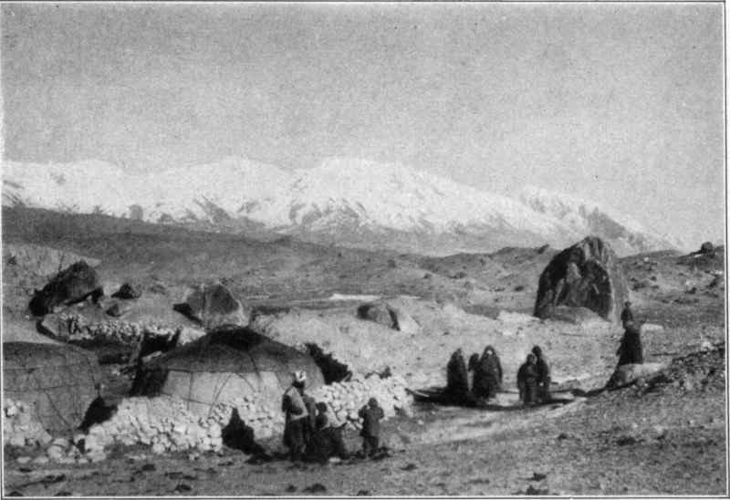 Two yurts in a barren landscape with people in the foreground and mountains in the background.